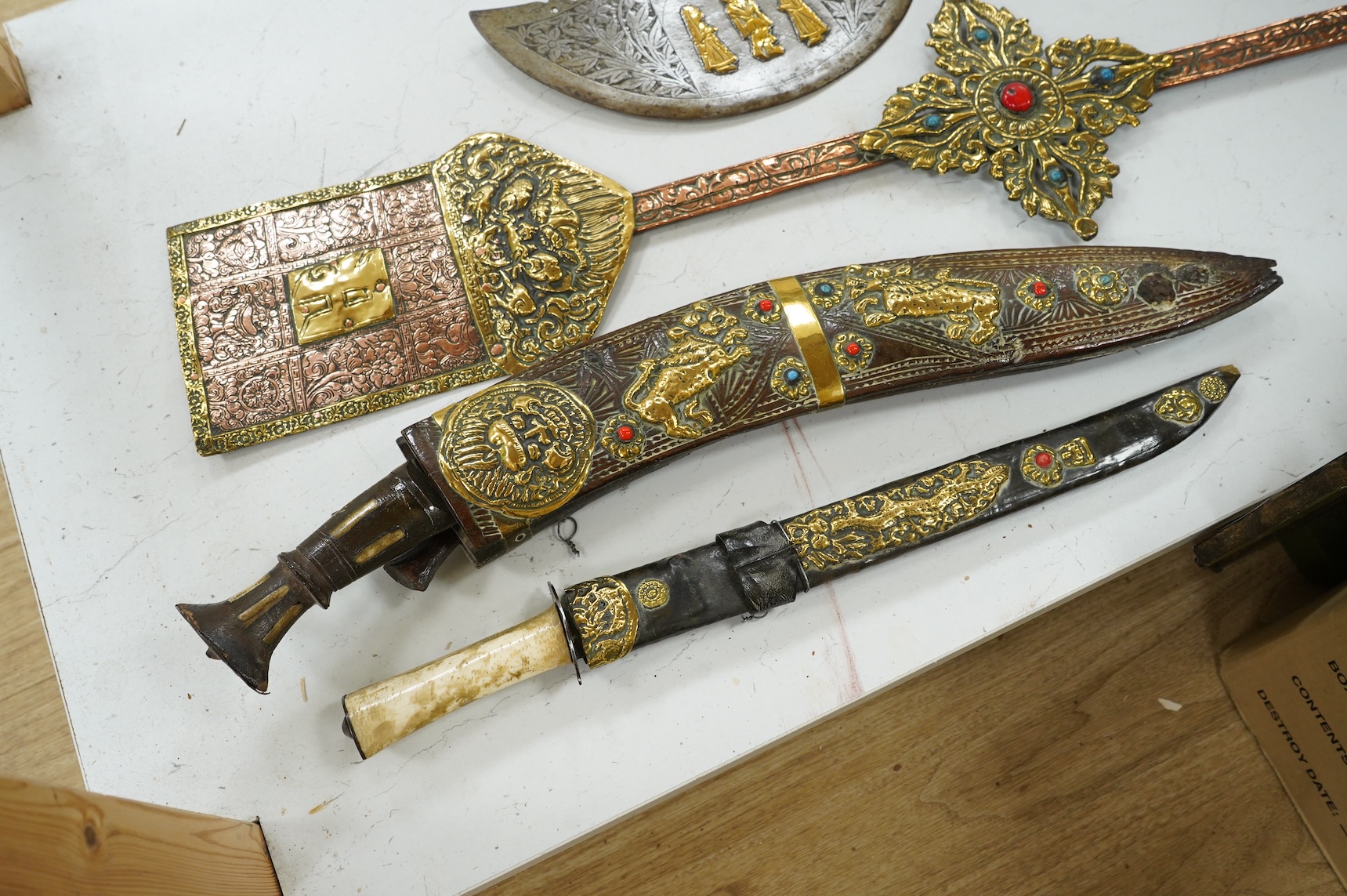 A decorative Indian axe, the large head inlaid with brass Hindu figures, together with a brass mounted North Indian kukri, another similar knife with bone handle, and a large shovel-shaped copper and brass item possibly